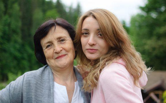 Close up portrait of happy smiling mother and daughter who are looking at the camera outside over landscape of forest and mountains.