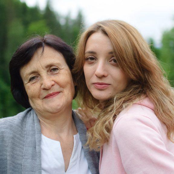 Close up portrait of happy smiling mother and daughter who are looking at the camera outside over landscape of forest and mountains.