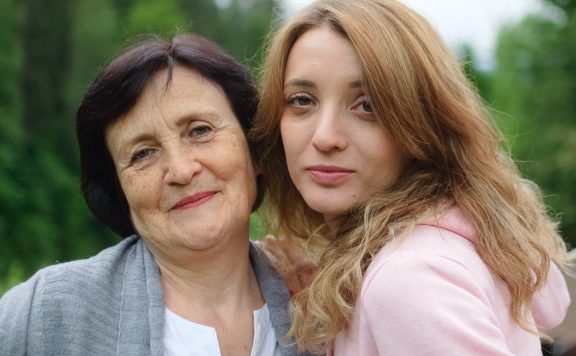 Close up portrait of happy smiling mother and daughter who are looking at the camera outside over landscape of forest and mountains.